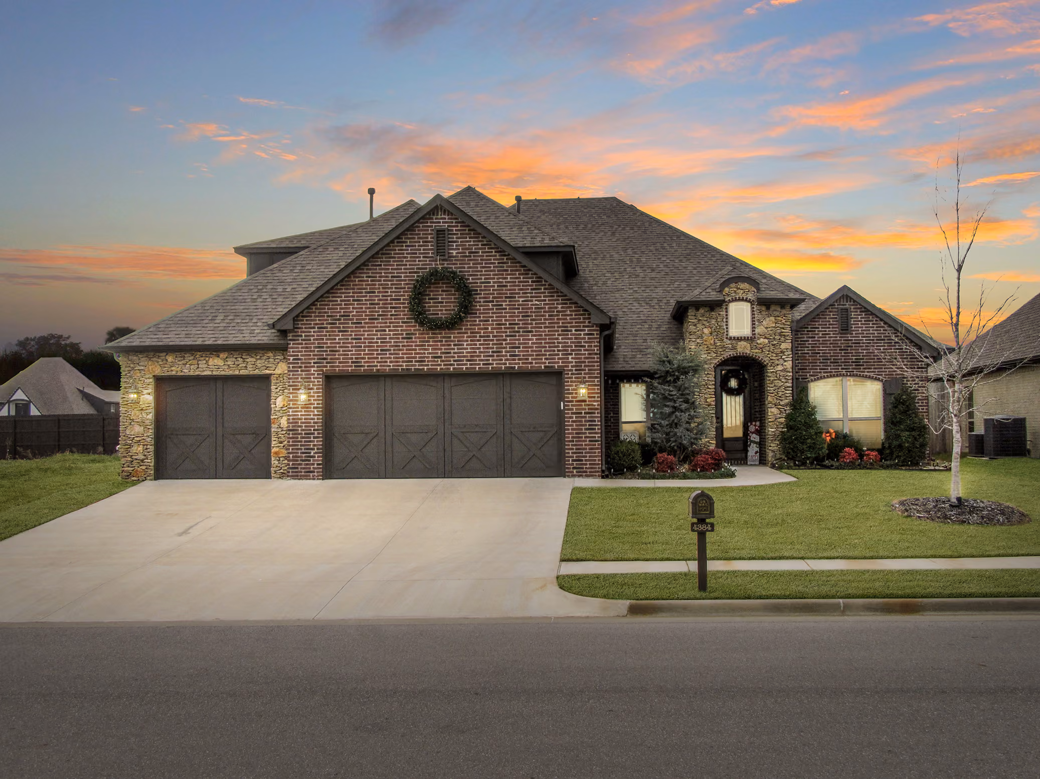Single-story brick and stone house with triple dark garage doors, a wreath above the center door, manicured lawn, and a mailbox in front at sunset.