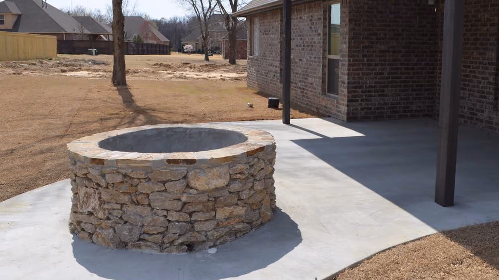 Stone fire pit on a concrete patio beside a brick house with a covered porch and a dry grassy yard.