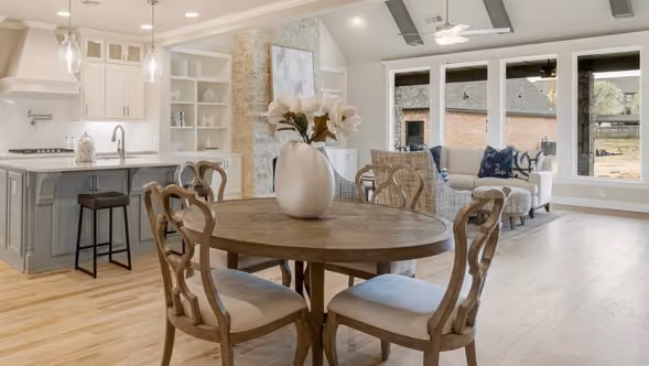 Bright open living and dining area with a round wooden table, cushioned chairs, a vase with flowers, and a modern kitchen with island and stools.