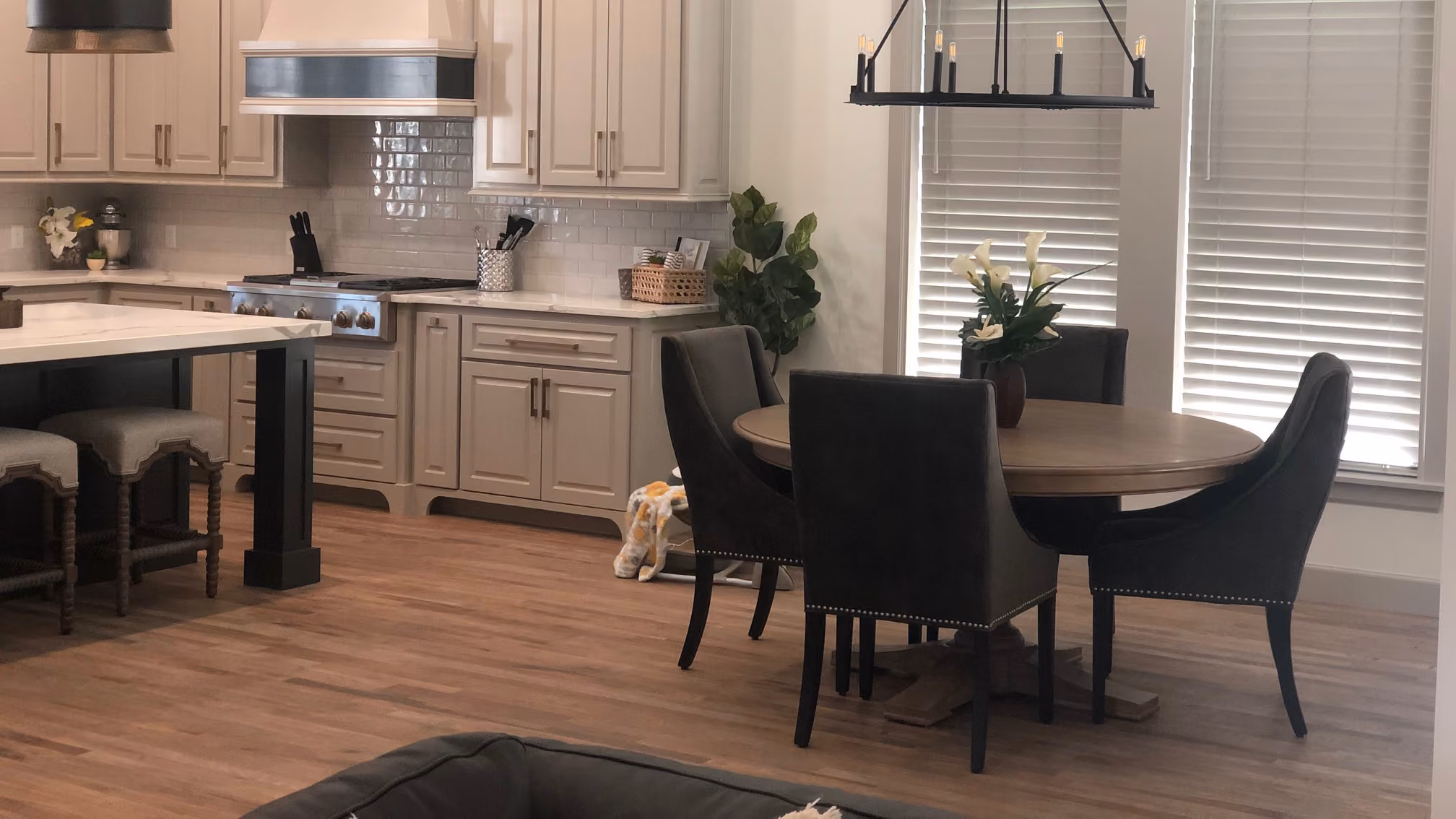 Modern kitchen and dining area with light wood flooring, beige cabinets, round wooden table, dark upholstered chairs, and large windows with blinds.