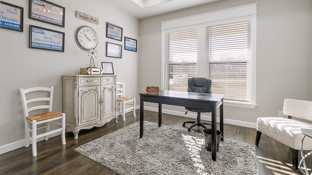 Bright office with a black desk, office chair, white rug, cabinet, two white chairs, and framed awards on a light gray wall.