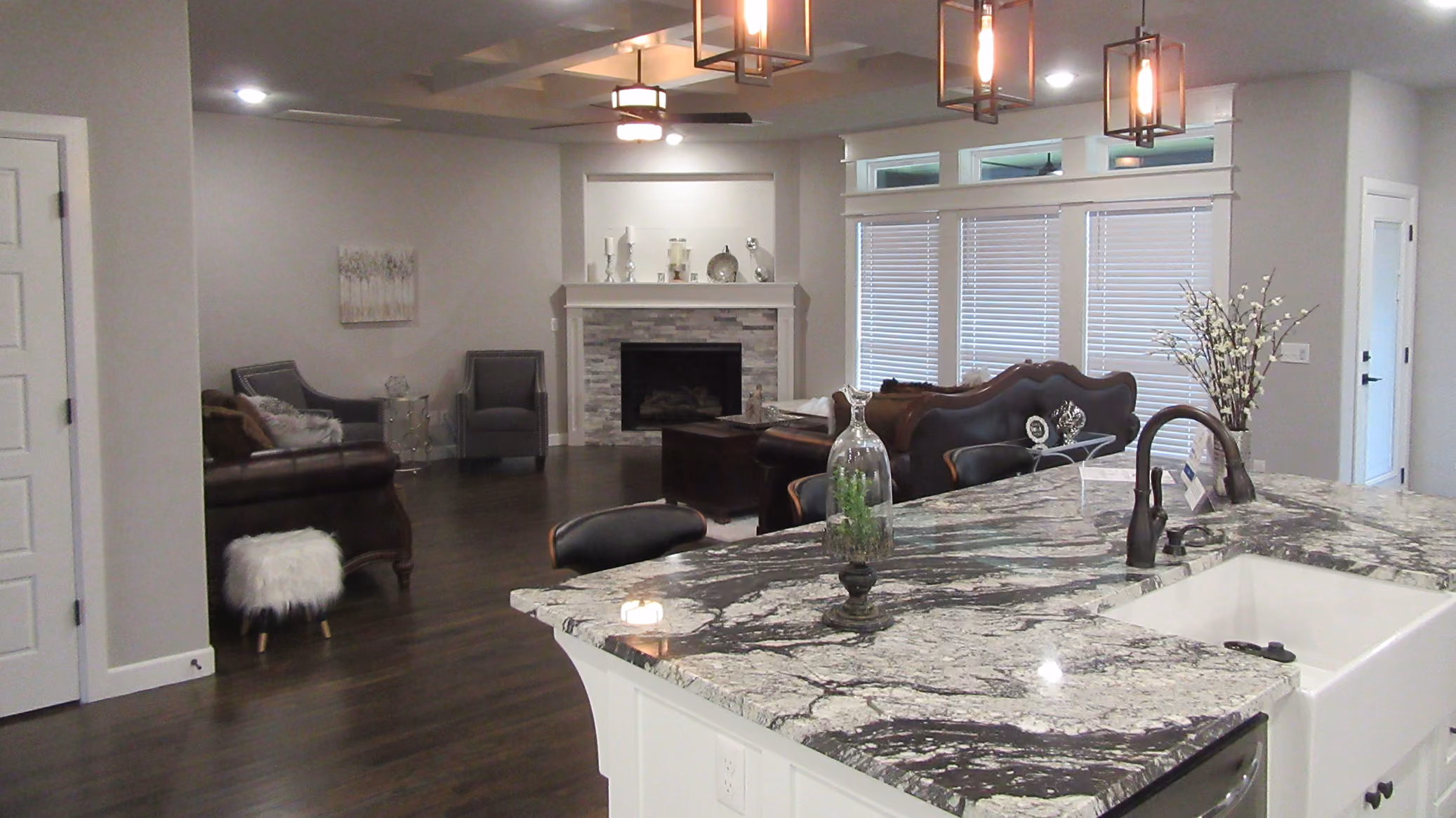 Modern open living space with dark wood floors, gray walls, a stone fireplace, leather sofa, and a kitchen island with black and white marble countertop and a sink.