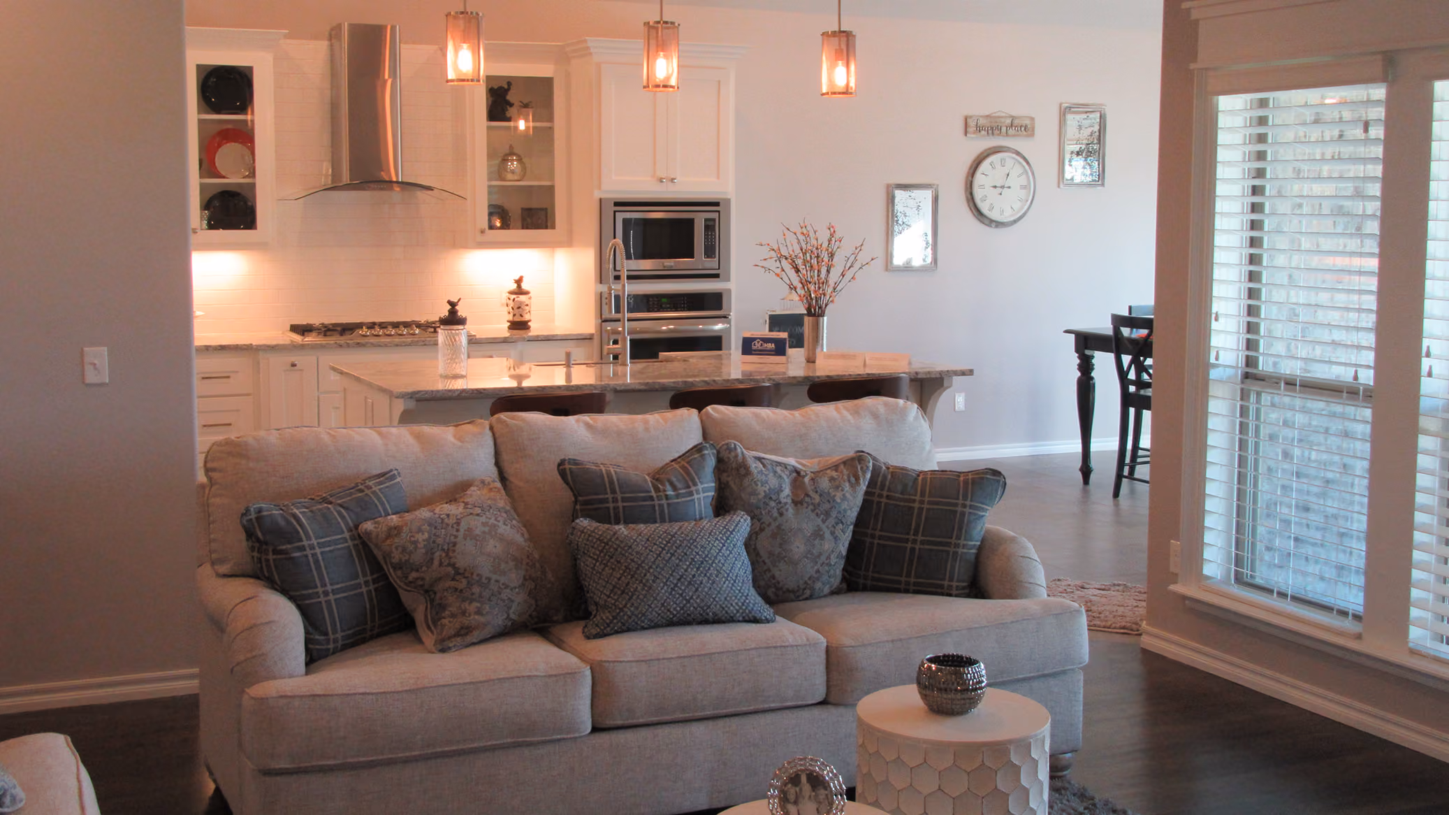 Modern living room with a beige sofa decorated with assorted patterned pillows, facing an open kitchen with a granite countertop island, stainless steel appliances, and pendant lights.