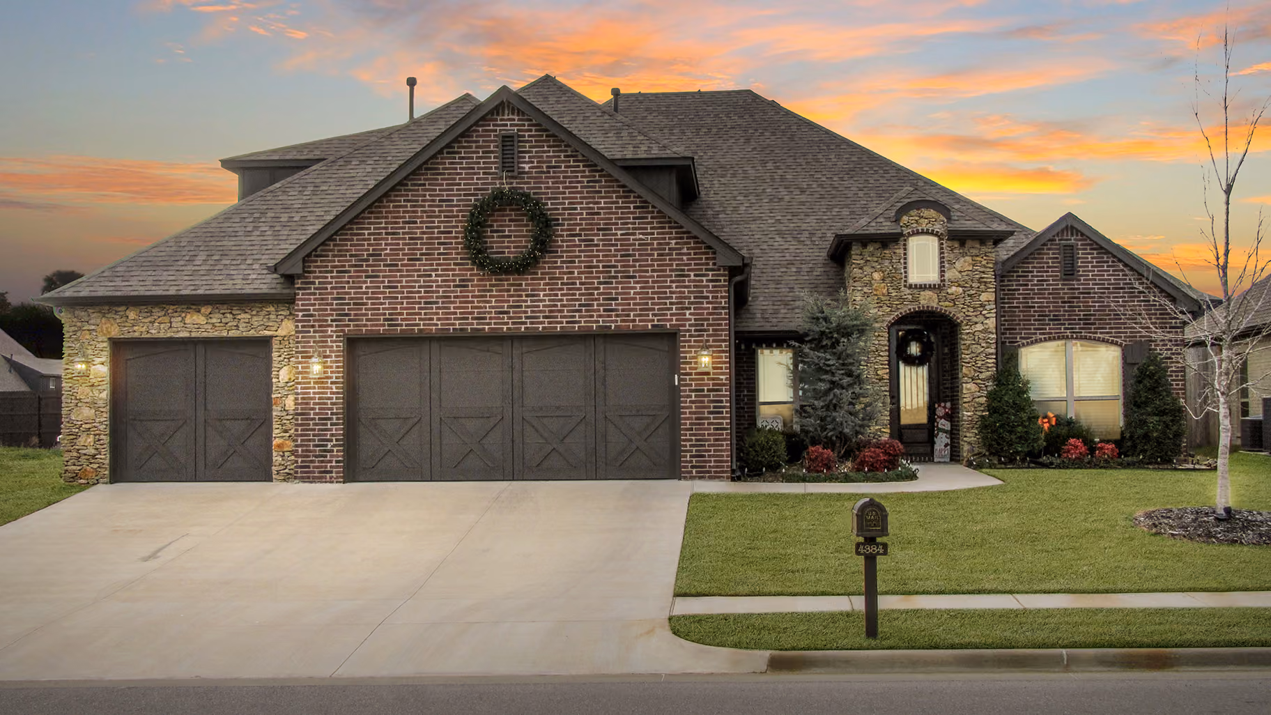 Single-story brick and stone house with a three-car garage, wreath decorations, and a well-maintained lawn at sunset.