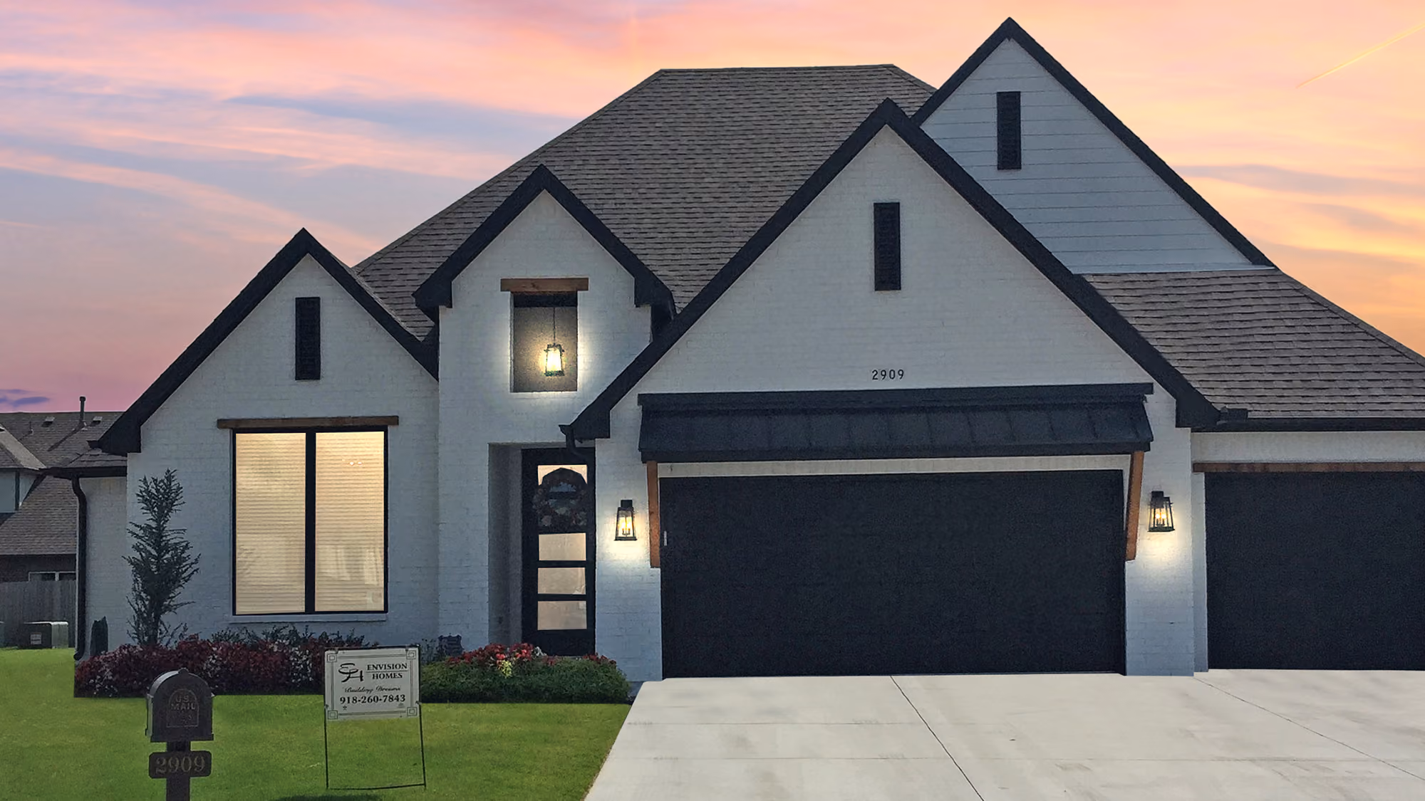 Modern white brick house with black trim, two-car garage, lit exterior sconces, and a sunset sky.