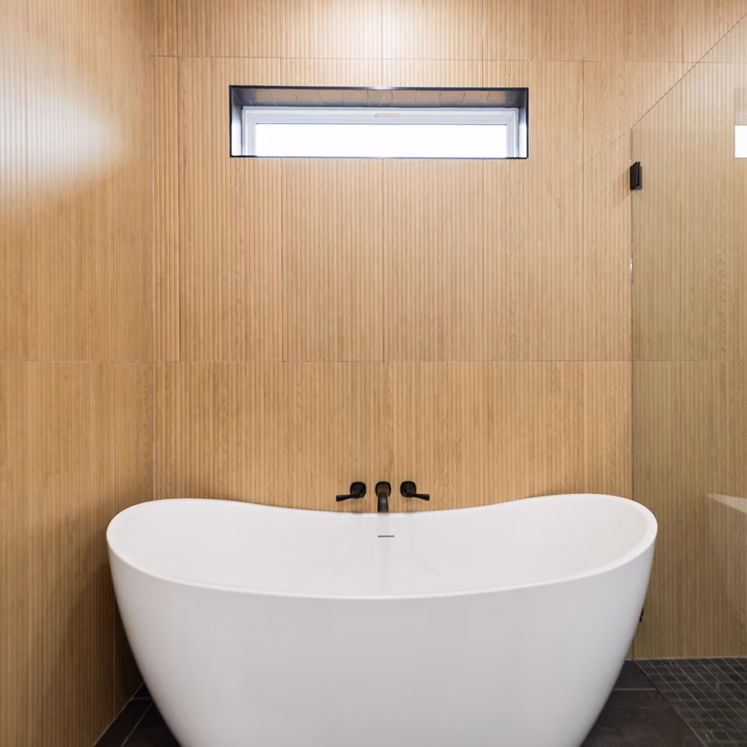 White freestanding bathtub against light wood paneling with a narrow window above and black fixtures.