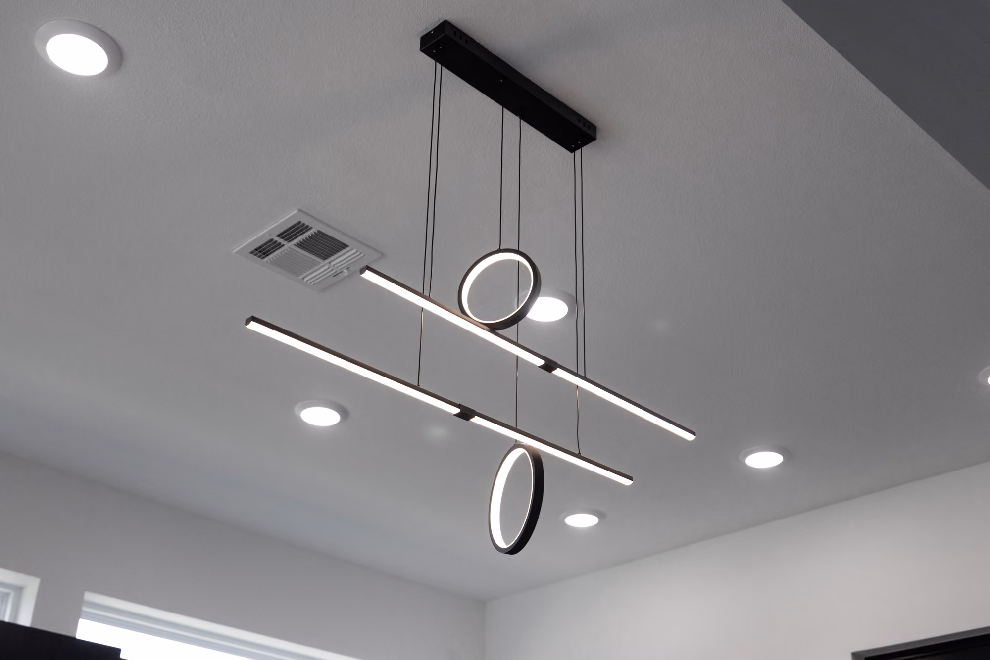 Modern ceiling light fixture with two horizontal bars and two illuminated circular rings hanging by wires.