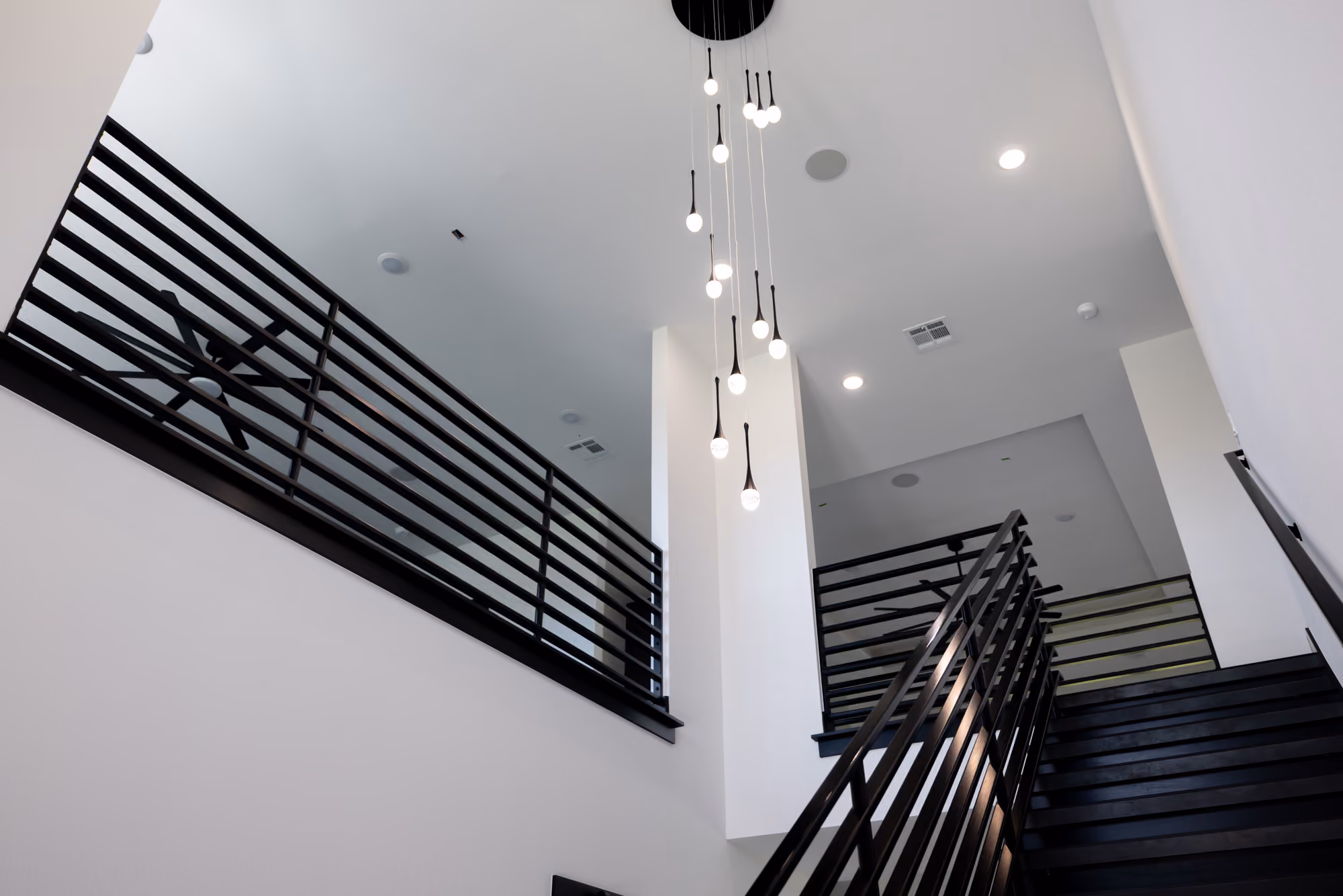Modern staircase with black railing, white walls, and hanging pendant lights from a high ceiling.