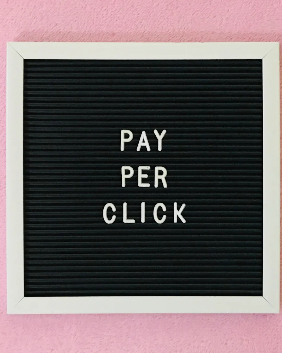 Black letter board with white frame displaying the words 'PAY PER CLICK' on a pink textured background.