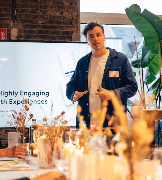 A man in a navy jacket presenting in front of a screen with blurred flowers and candles in the foreground.