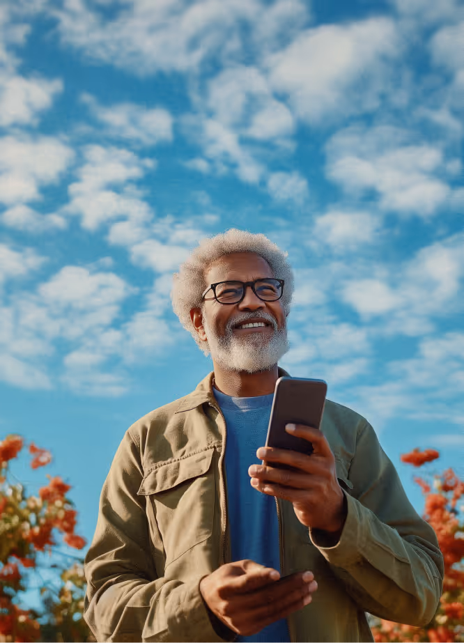 Smiling elderly man with gray hair and beard holding a smartphone outdoors under a partly cloudy blue sky.