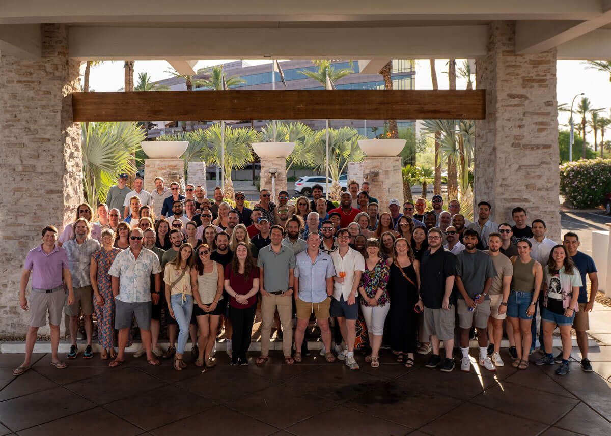 Intellum company photo of a large group of diverse people standing together outdoors under a shaded archway with palm trees and modern buildings in the background.