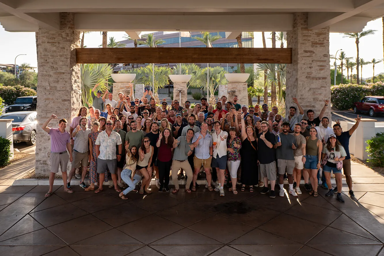 Large group of diverse people posing happily under a stone and wood pergola outside, many waving or making hand signs.