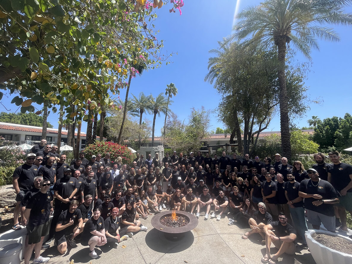 Large group of people wearing black shirts posing outdoors around a fire pit with palm trees and blue sky in the background.
