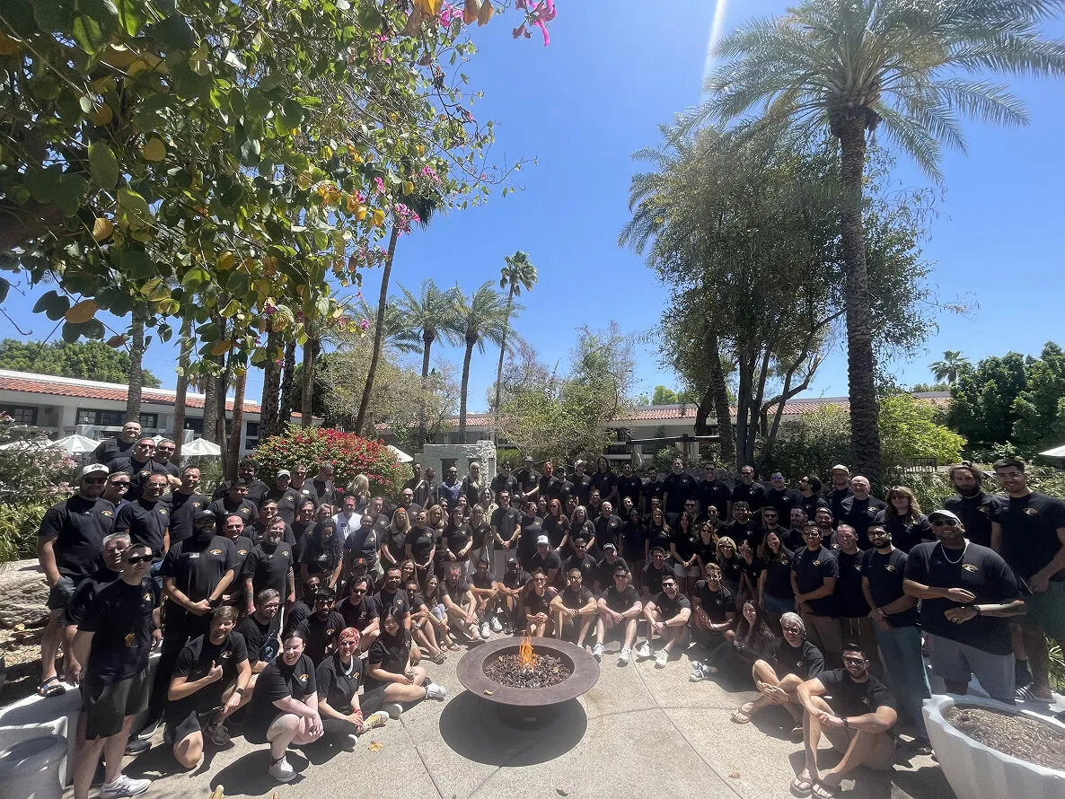 Large group of people wearing black shirts posing outdoors around a fire pit with palm trees and blue sky in the background.