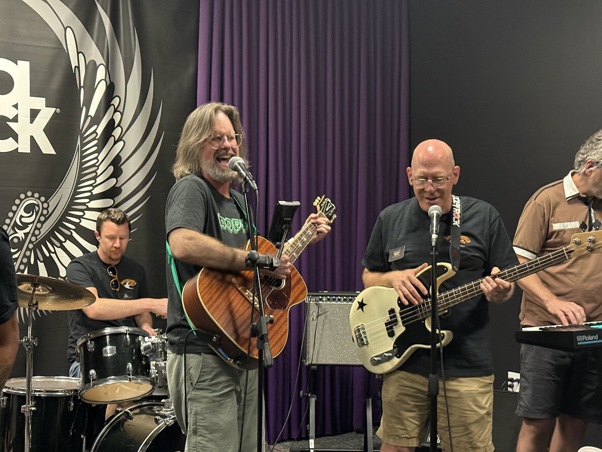 Four men playing music in a rehearsal room: one on drums, one singing and playing acoustic guitar, one singing and playing bass guitar, and one playing keyboard.