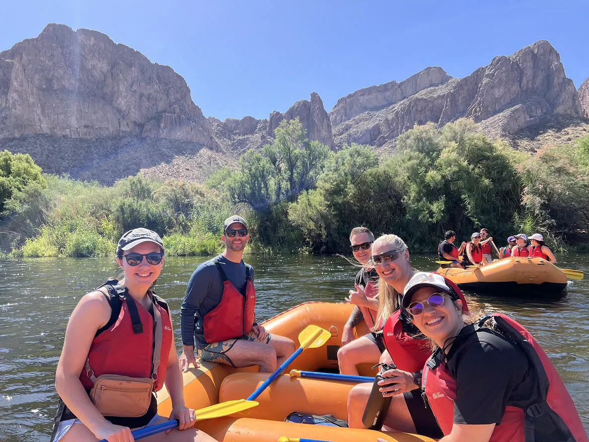 Group of people wearing red life jackets and sunglasses rafting on a river with rocky mountains and green bushes in the background.