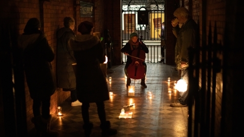 We are in a dark alley with red brick walls and black and white tiles. A young woman sits wrapped in winter clothes playing the cello surrounded by candles which glisten on the tiled floor. A small socially distanced audience watch her play. At the other end of the alley is an iron gate and a figure can be seen watching through the gate