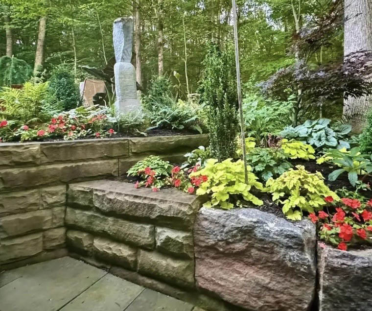 Akron garden on a sunny day, with a retaining wall, tall stone statue, and many green plants and red flowers