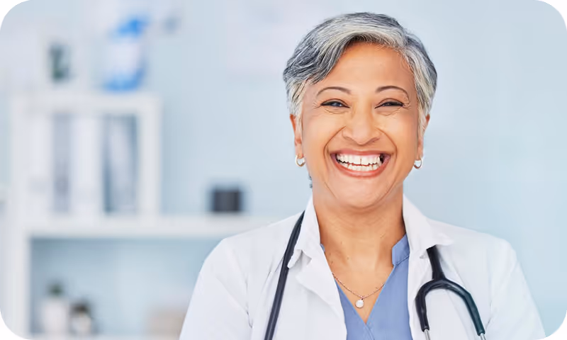 Smiling female doctor with short gray hair wearing a white coat and stethoscope in a medical office.