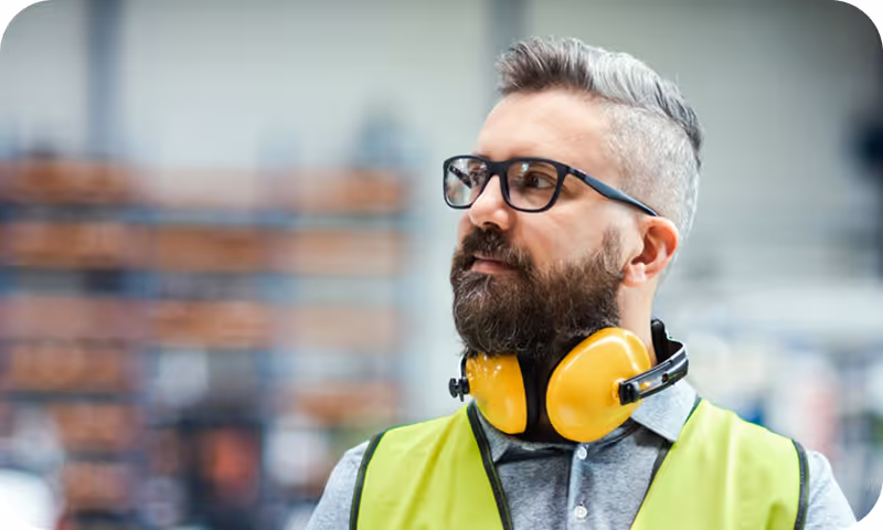 Man with glasses and gray hair wearing a yellow safety vest and yellow ear protection in an industrial setting.