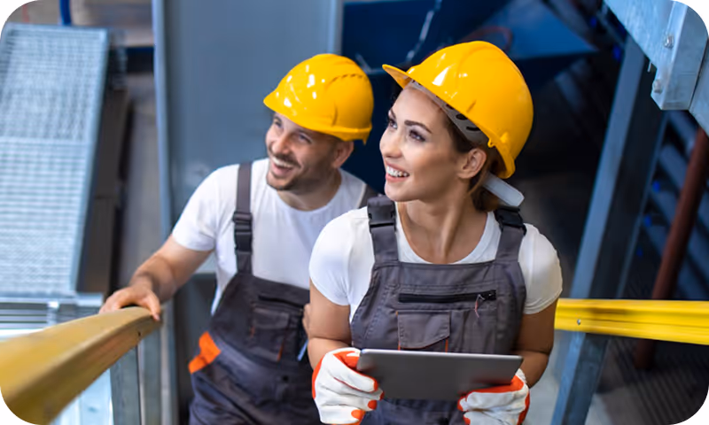Two construction workers in yellow hard hats and gray overalls smiling while holding a tablet and standing on stairs with yellow handrails.