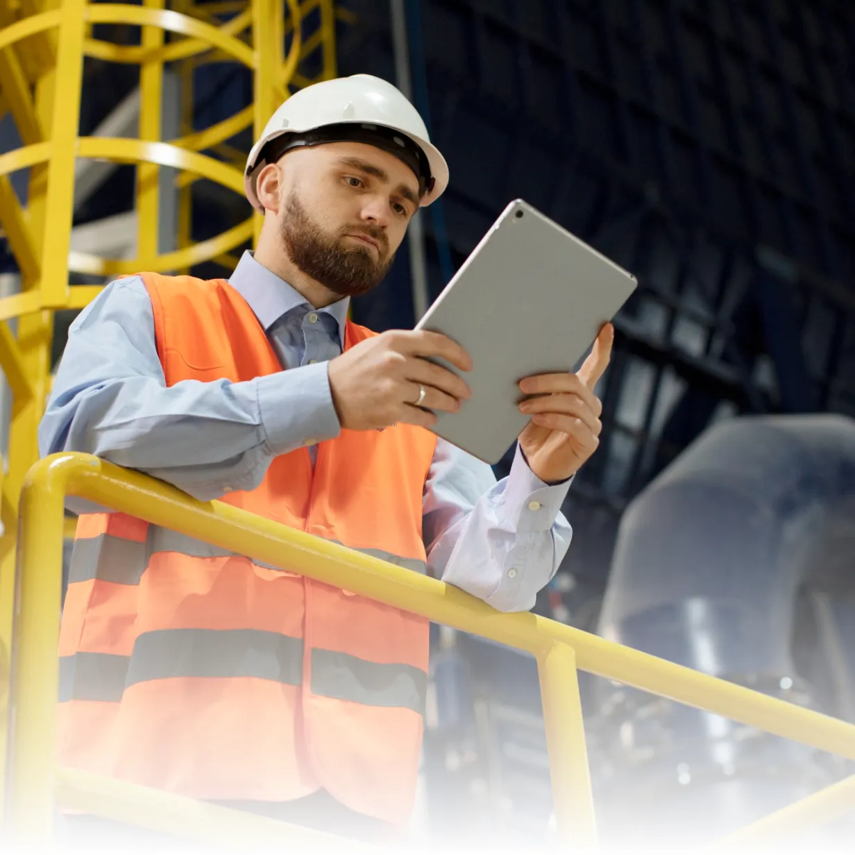 Ingenieur mit Schutzhelm und Warnweste liest Tablet auf gelbem Laufsteg in Industriehalle