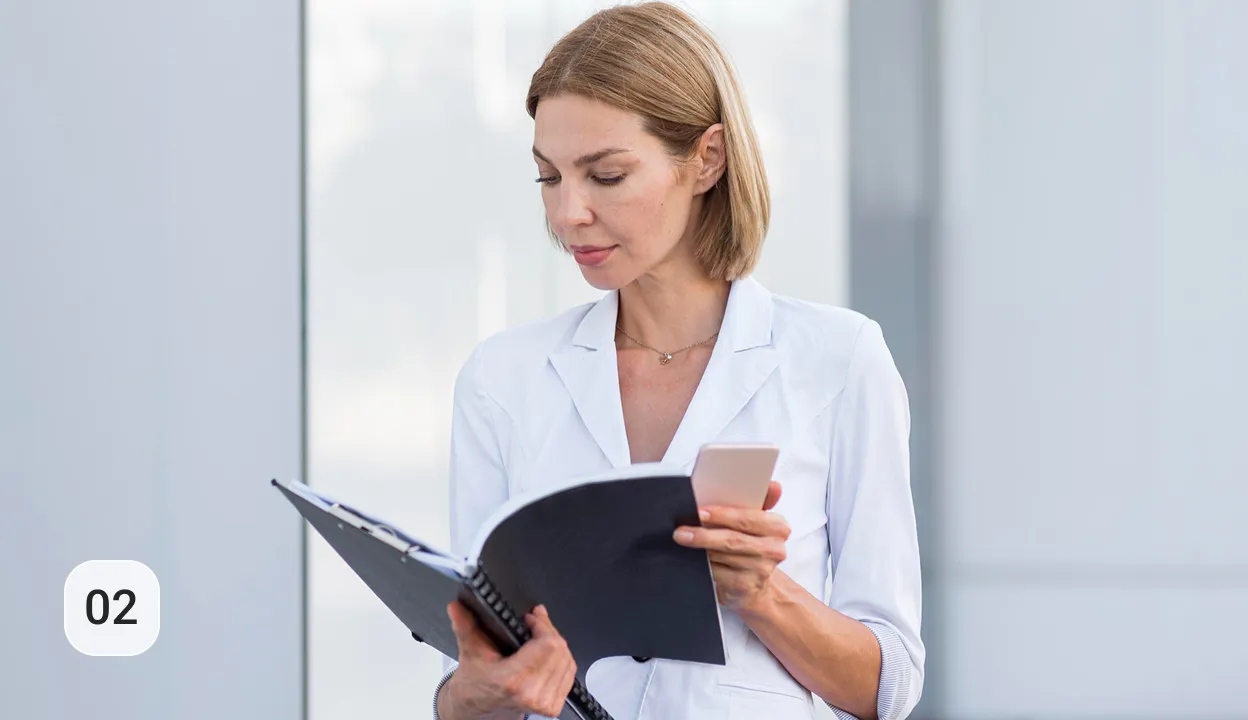 Ältere Frau in weißem Blazer liest in einem Notizbuch und hält ein Smartphone, stehende Aufnahme vor hellem Hintergrund