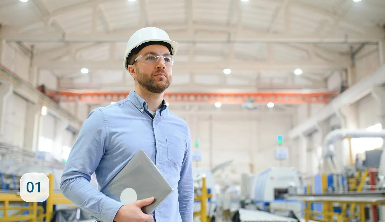 Ingenieur mit Schutzhelm und -brille hält Tablet in moderner, hell beleuchteter Industriehalle mit Maschinen im Hintergrund