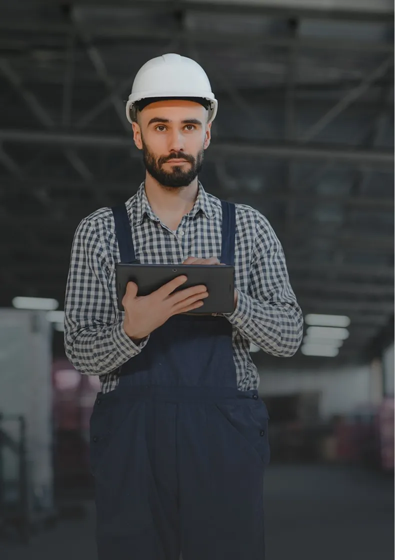 Mann mit Helm und Latzhose hält Tablet in Industriehalle, ernster Blick, Hintergrund verschwommen