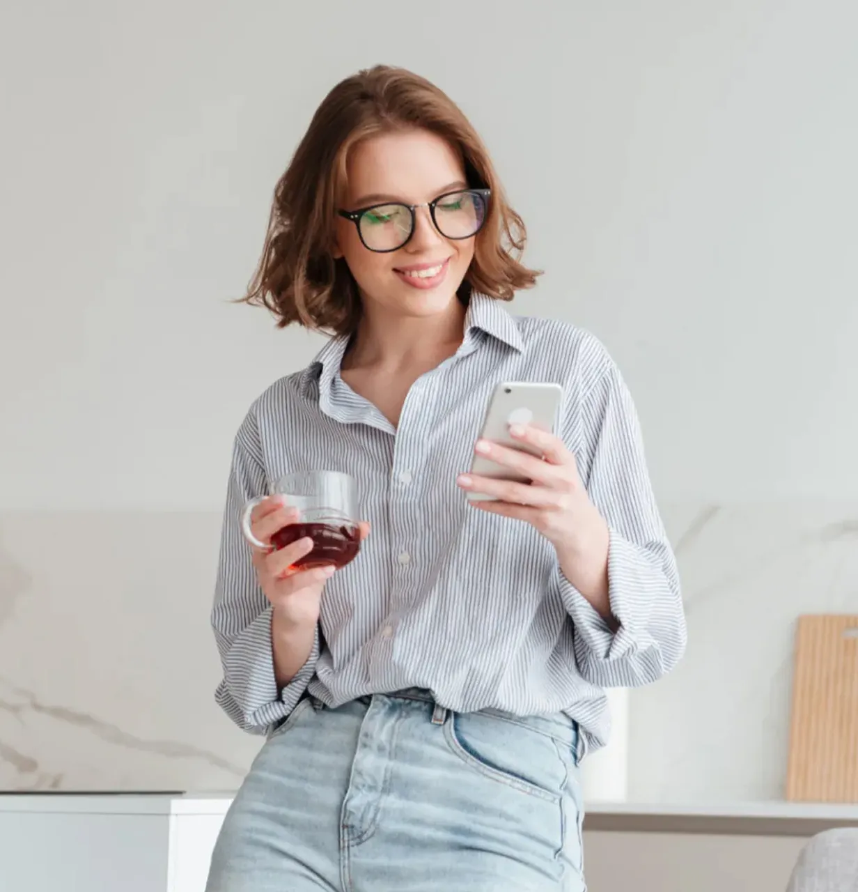 junge Frau mit Brille liest auf Smartphone, hält Tasse Tee, lächelnd in lockerer Bluse und Jeans in heller Küche