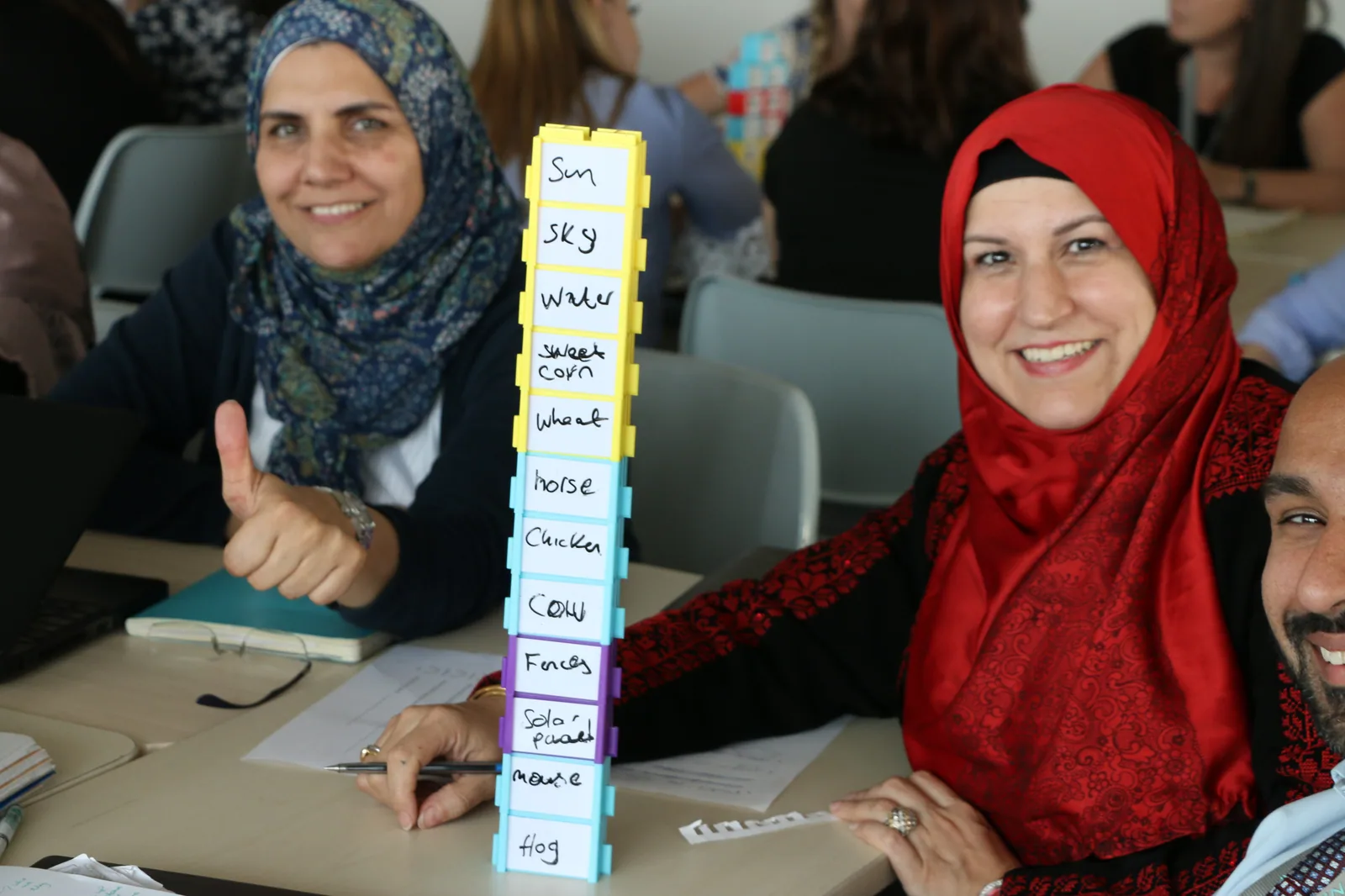Two teachers smiling beside a tower of Writer's Block they have labelled sun, sky, water, wheat, horse, chicken, cow and frog.