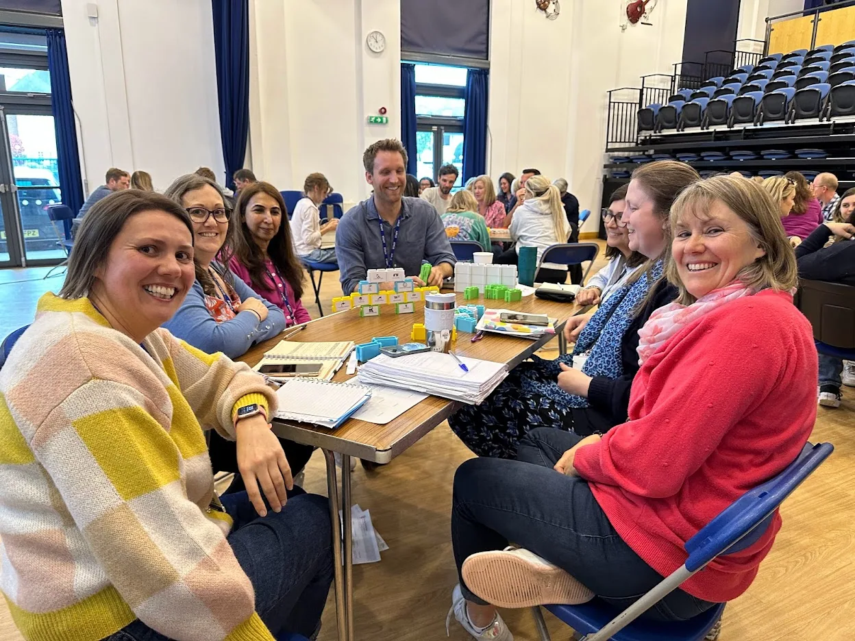 Teachers at round tables in a school hall, smiling at the camera during a staff session.