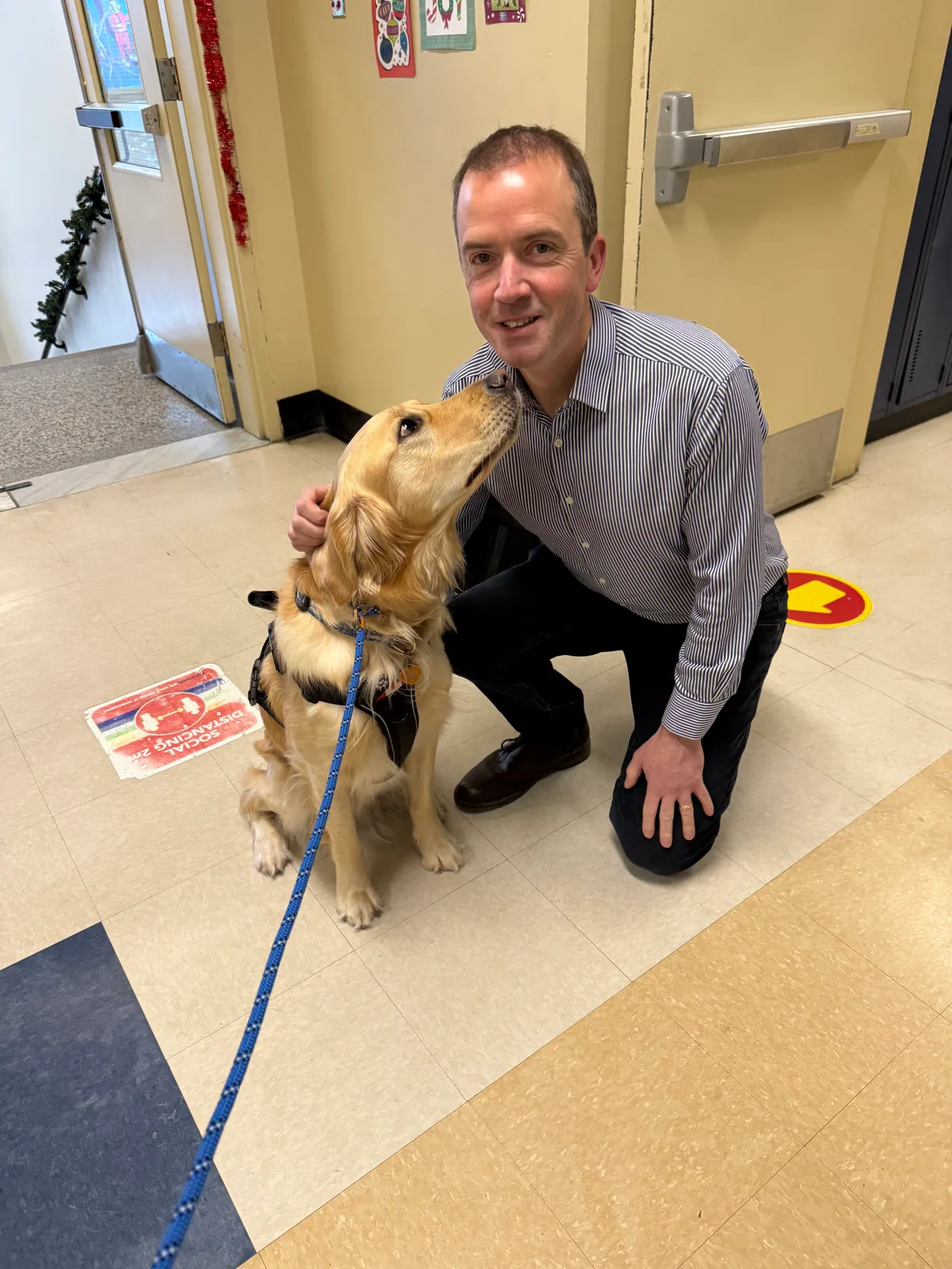 Paul Main kneeling in a school corridor in Quebec, laughing as the school dog, a golden retriever, licks his face