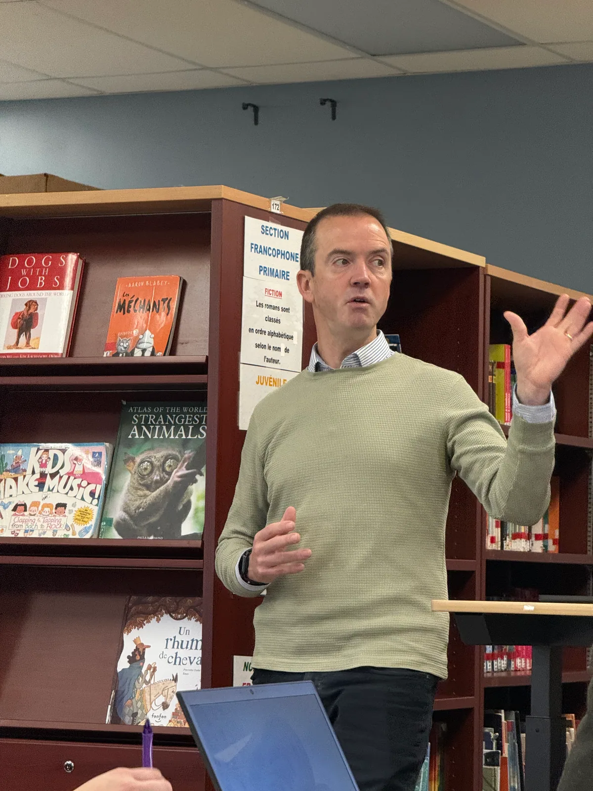 Paul Main mid-sentence, gesturing, in front of the shelves of a school library in Quebec