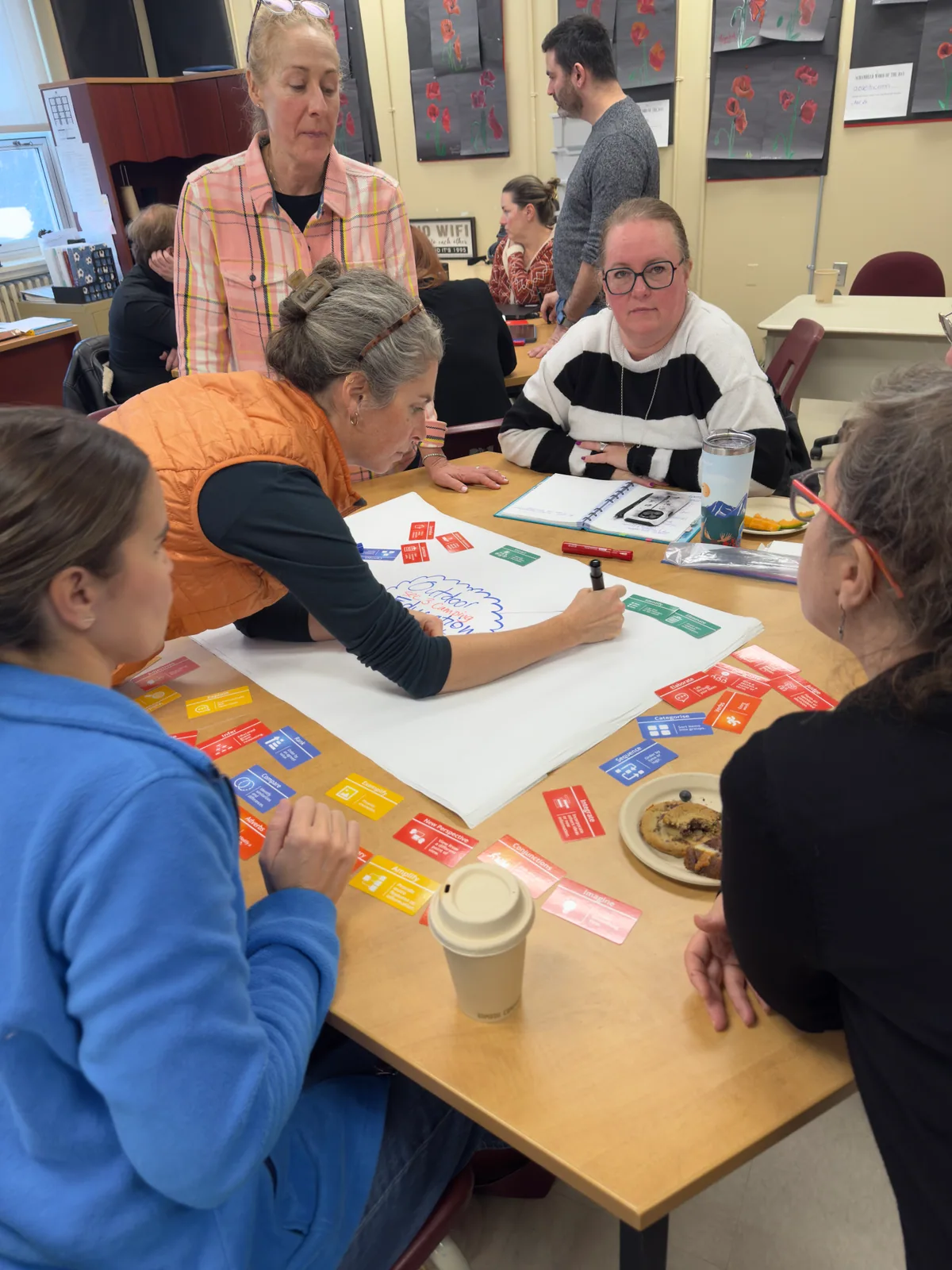 A teacher in Quebec leans over a large sheet, drawing a plan with Thinking Framework cards laid across it, colleagues working round the table.