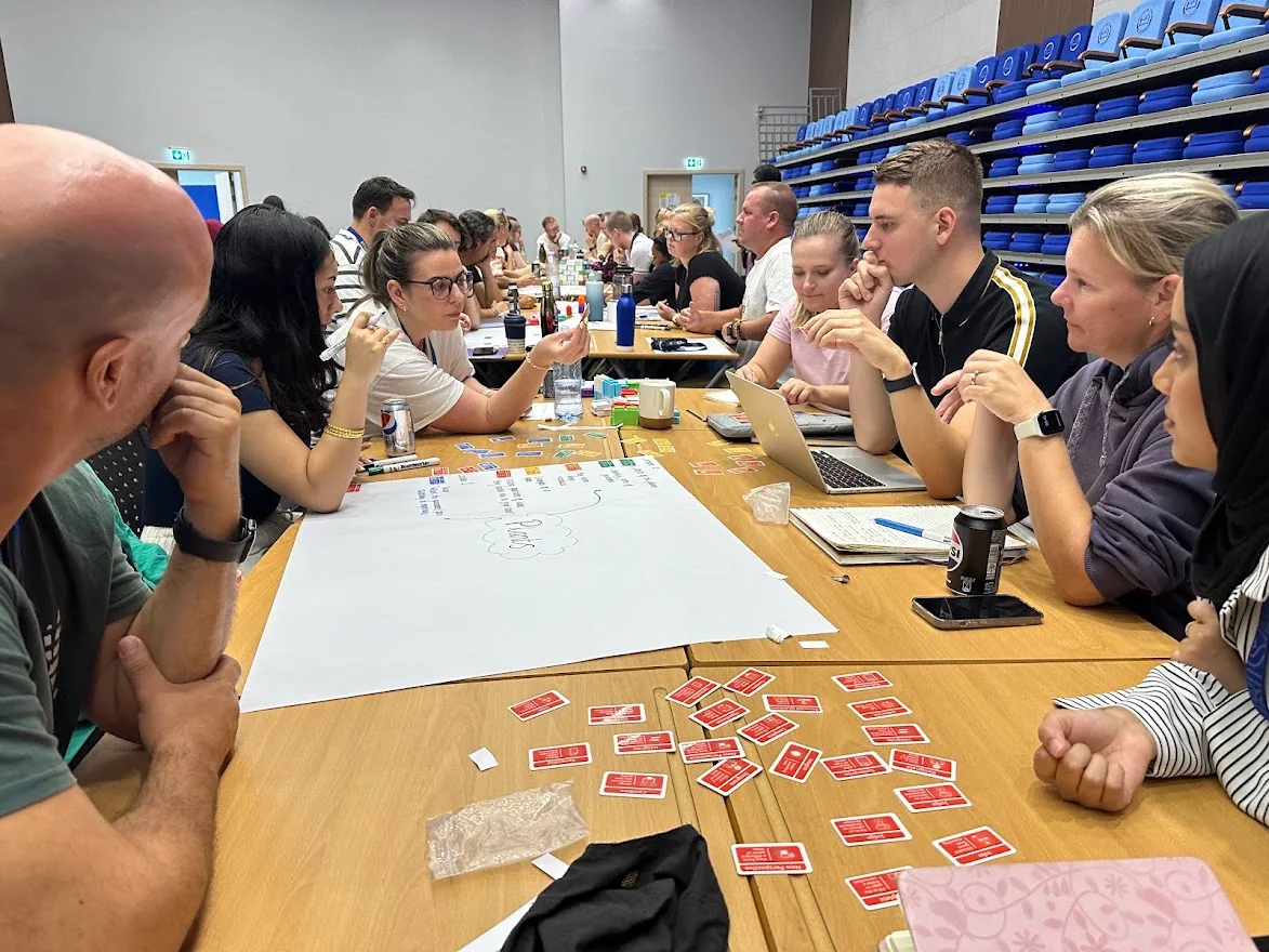 A long table of teachers leaning over a large sheet of paper, red Thinking Framework cards spread across the table, one teacher pointing at the plan