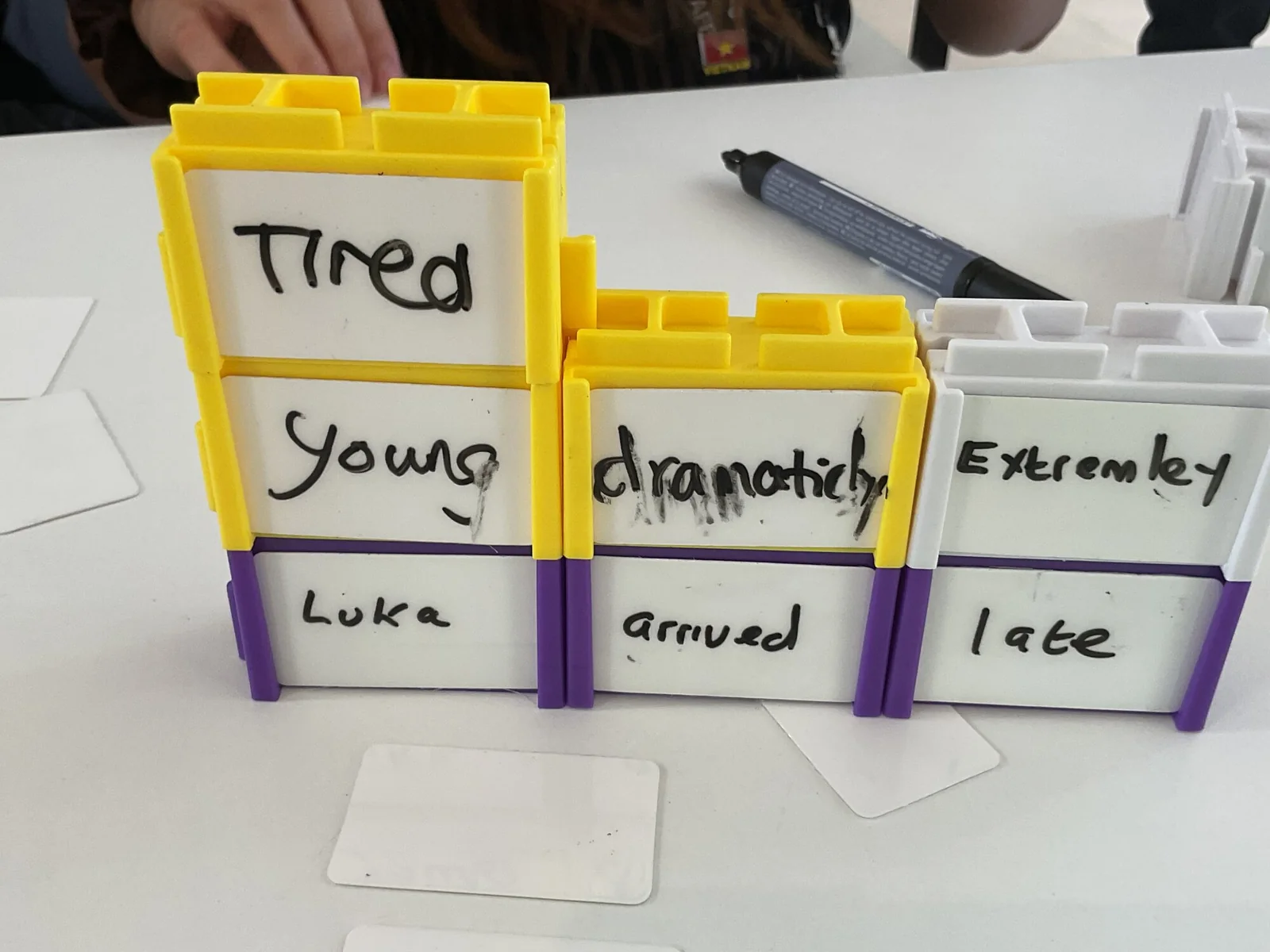 A sentence built on a desk from blocks: purple blocks read Luka, arrived, late, with yellow and white blocks stacked on top reading Tired, Young, dramatically and Extremely, handwritten in dry-wipe pen