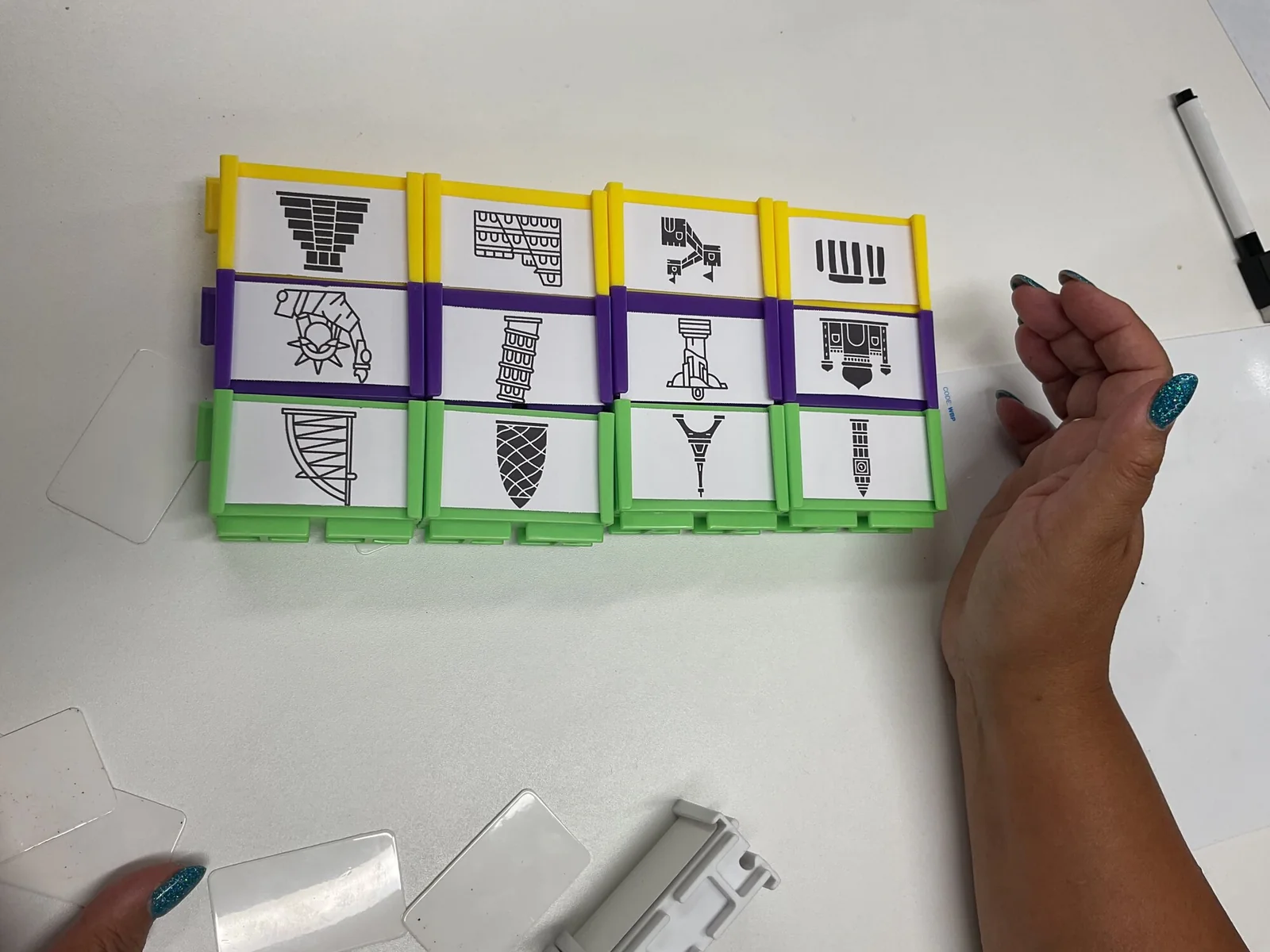 A grid of twelve blocks in yellow, purple and green, each holding a printed line drawing of a famous landmark, with a hand resting beside it and spare mini whiteboards on the table