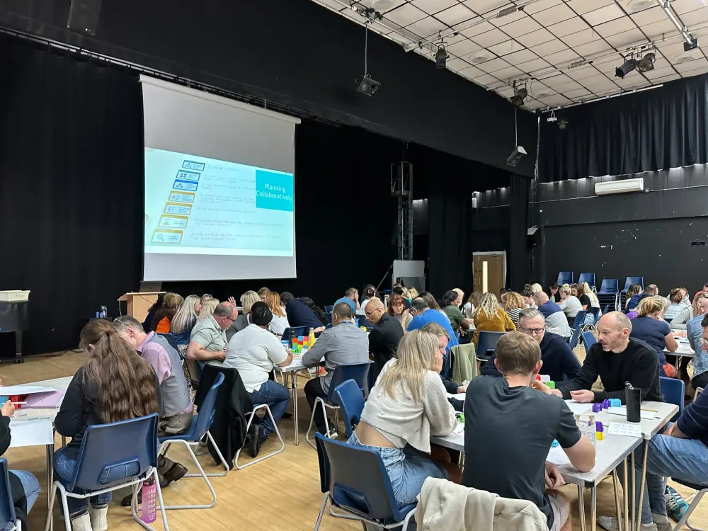 A school hall full of teachers at long tables, seen from the back, a Thinking Framework slide on the screen at the front