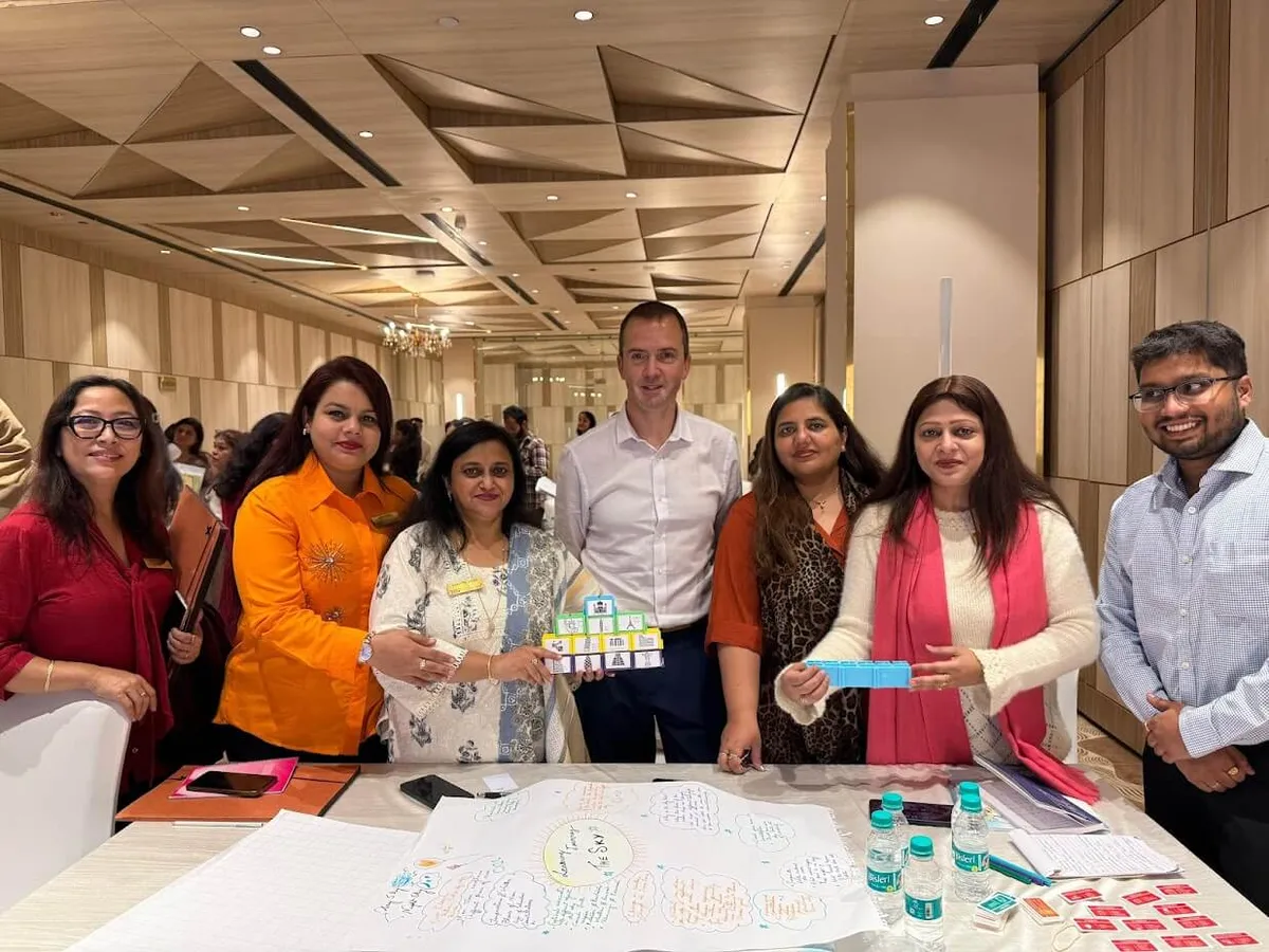 Paul Main standing with a teaching team at a conference table, one holding a small tower of labelled blocks, a drawn plan on the table in front of them.