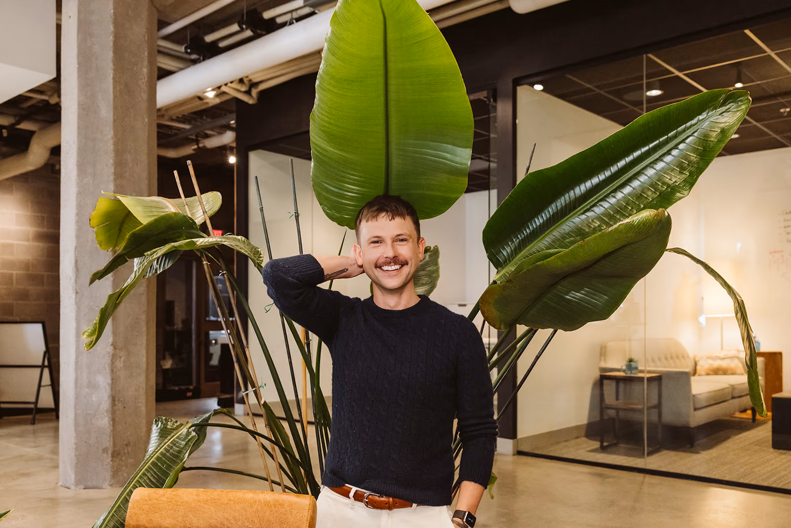 Man with a mustache wearing a black sweater and white pants smiling and posing with his hand behind his head in front of large green tropical leaves indoors near a glass-walled office space.