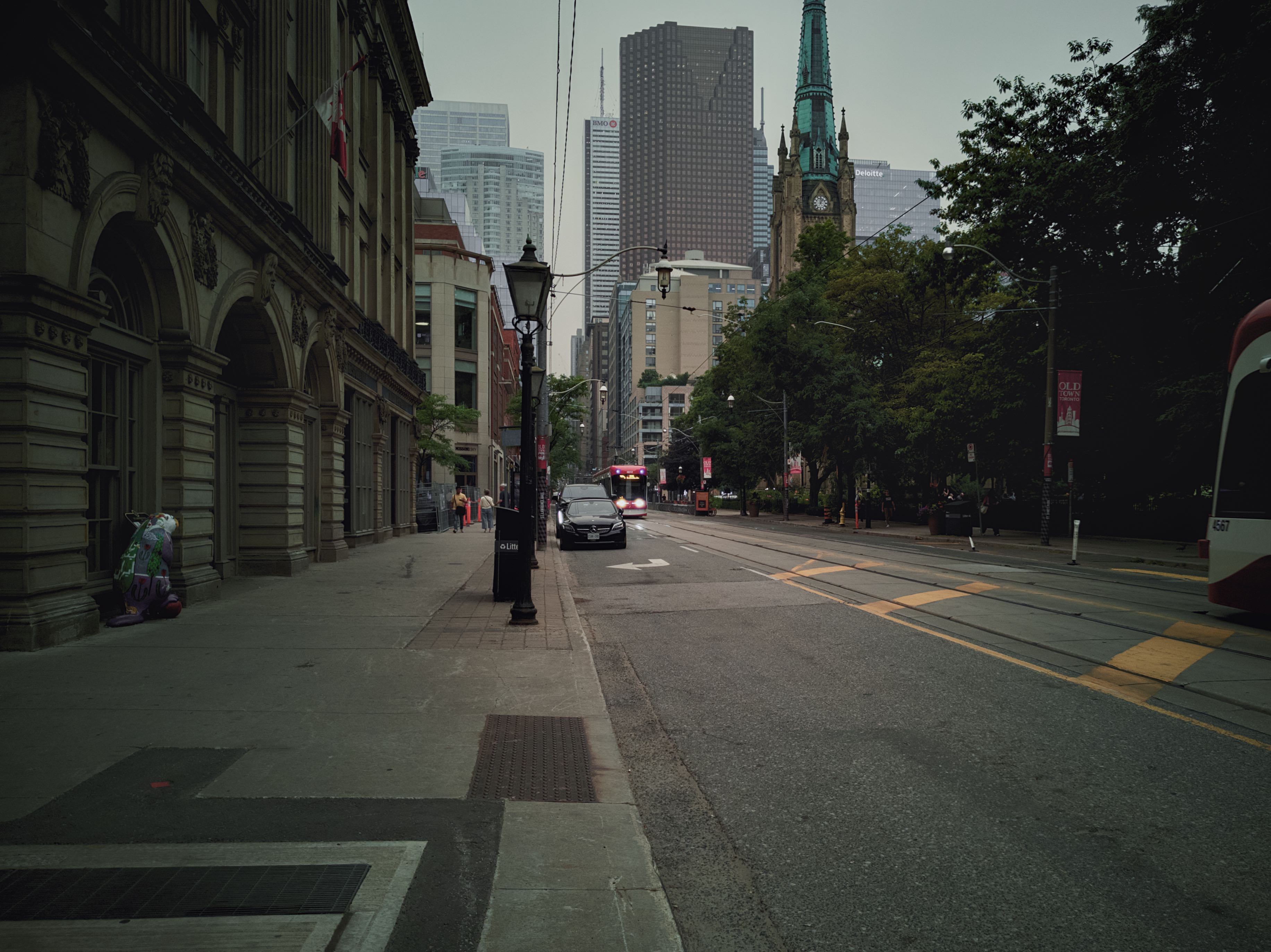 A street scene in a historic district during midday, featuring ornate, older buildings lining the sidewalk, a street with traffic, and lush green trees lining the road, with hazy, smoke-filled air.