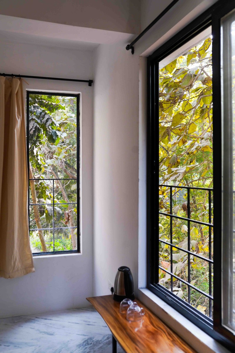 Room corner with two windows showing green trees and a wooden table with a kettle and glasses.