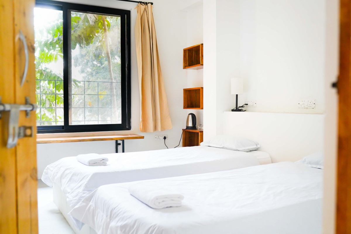 Bright room with two single beds, white linens, a window with beige curtains, and a wooden shelf with a kettle.