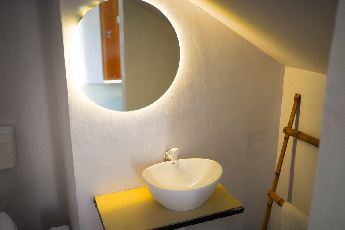 Round mirror with backlight above a white sink in a small bathroom with towel rack.