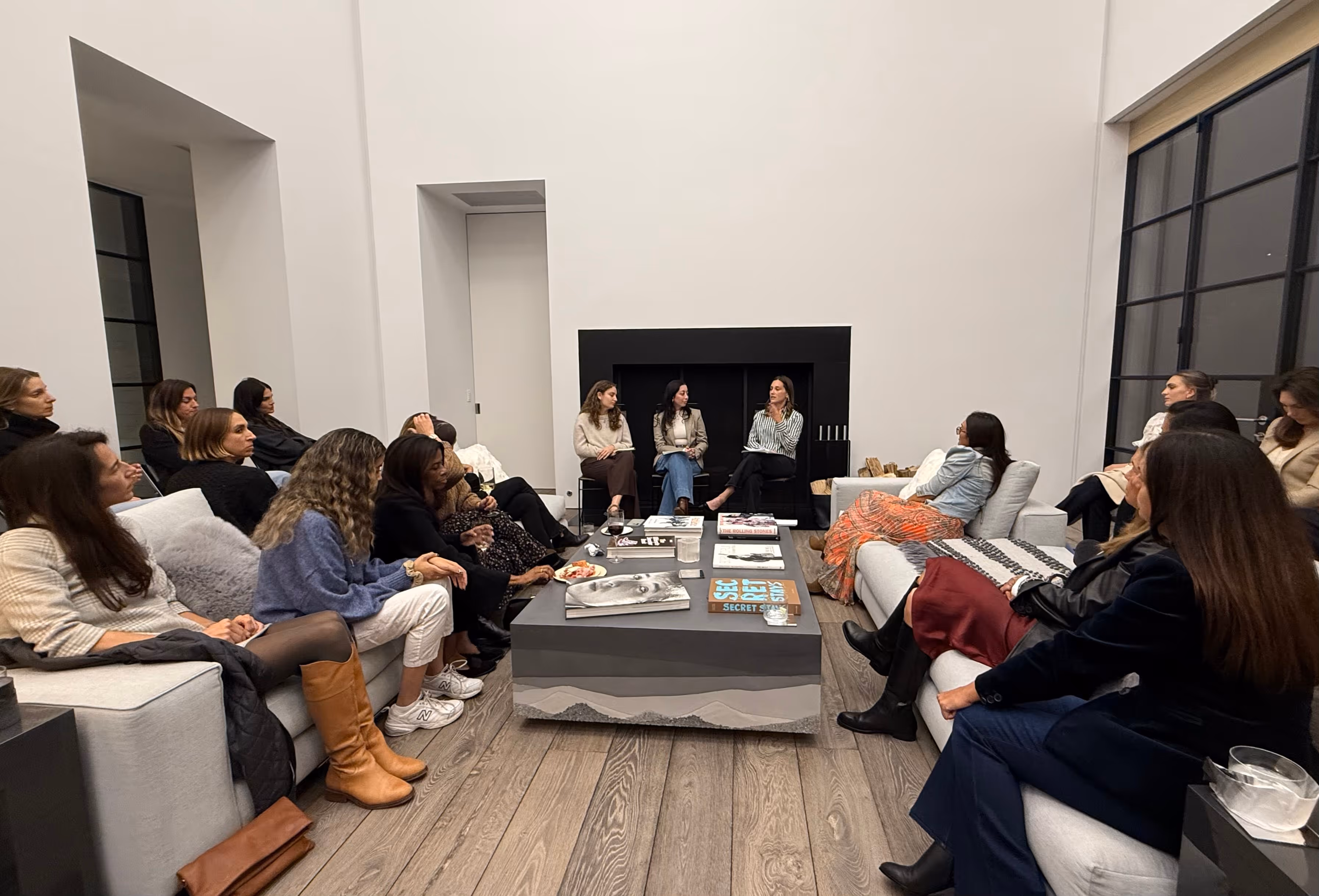 Group of women gathered in a modern living room engaged in a discussion around a coffee table with books and drinks.