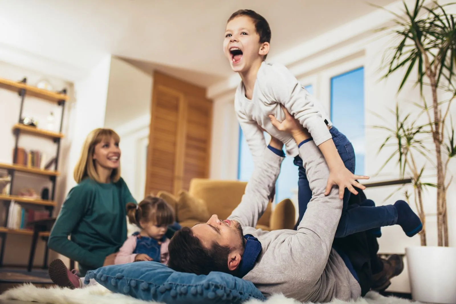 Father lying on the floor lifting his laughing son in the air while mother and daughter watch smiling in a cozy living room.