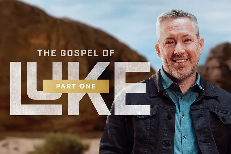 Smiling man in a black jacket and blue shirt standing outdoors with text 'The Gospel of Luke Part One'.