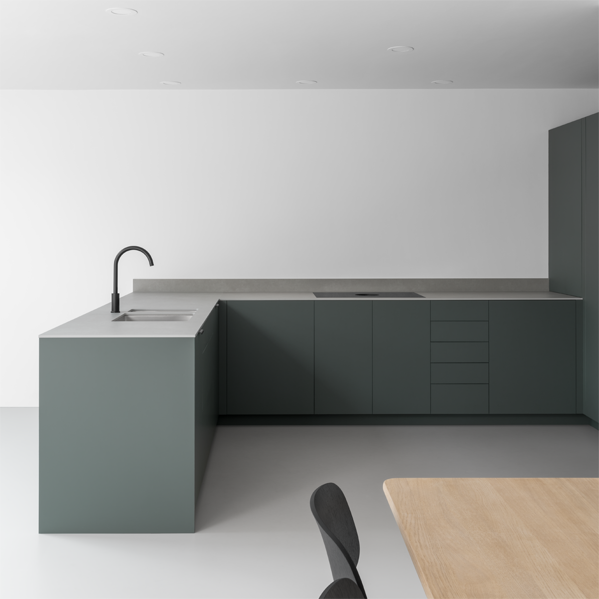 Minimalist architectural reconfiguration featuring open-plan kitchen dining room with grey resin floor, white walls, green grey cabinets and oak table.